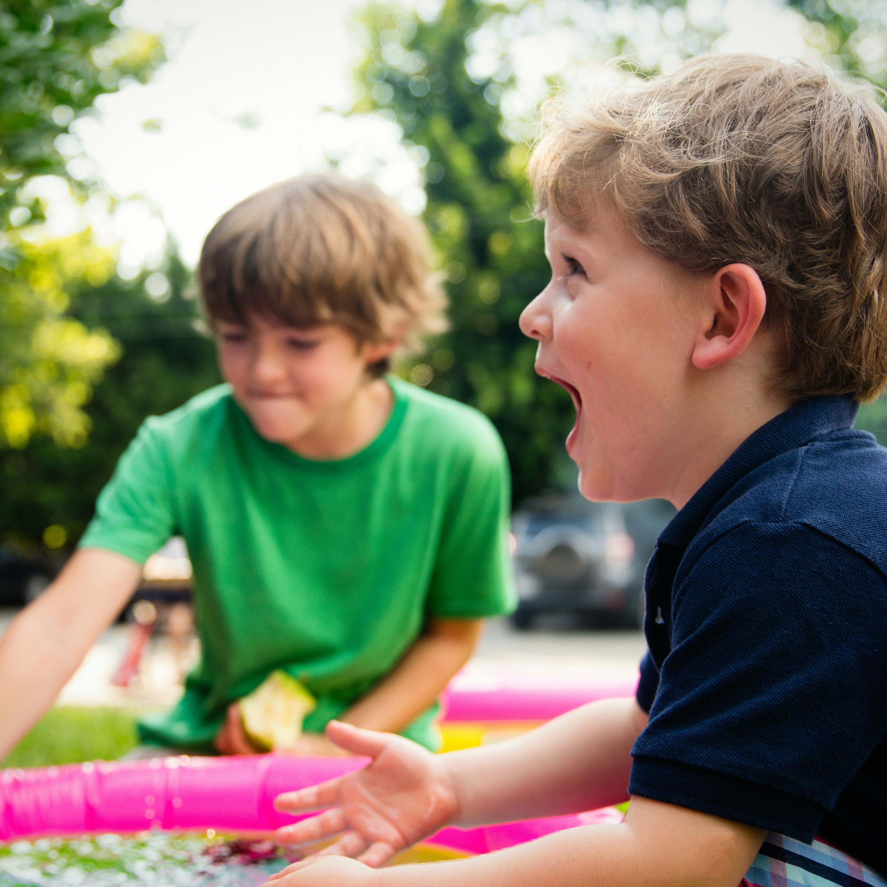 Two young boys playing and laughing near a small inflatable pool outdoors.