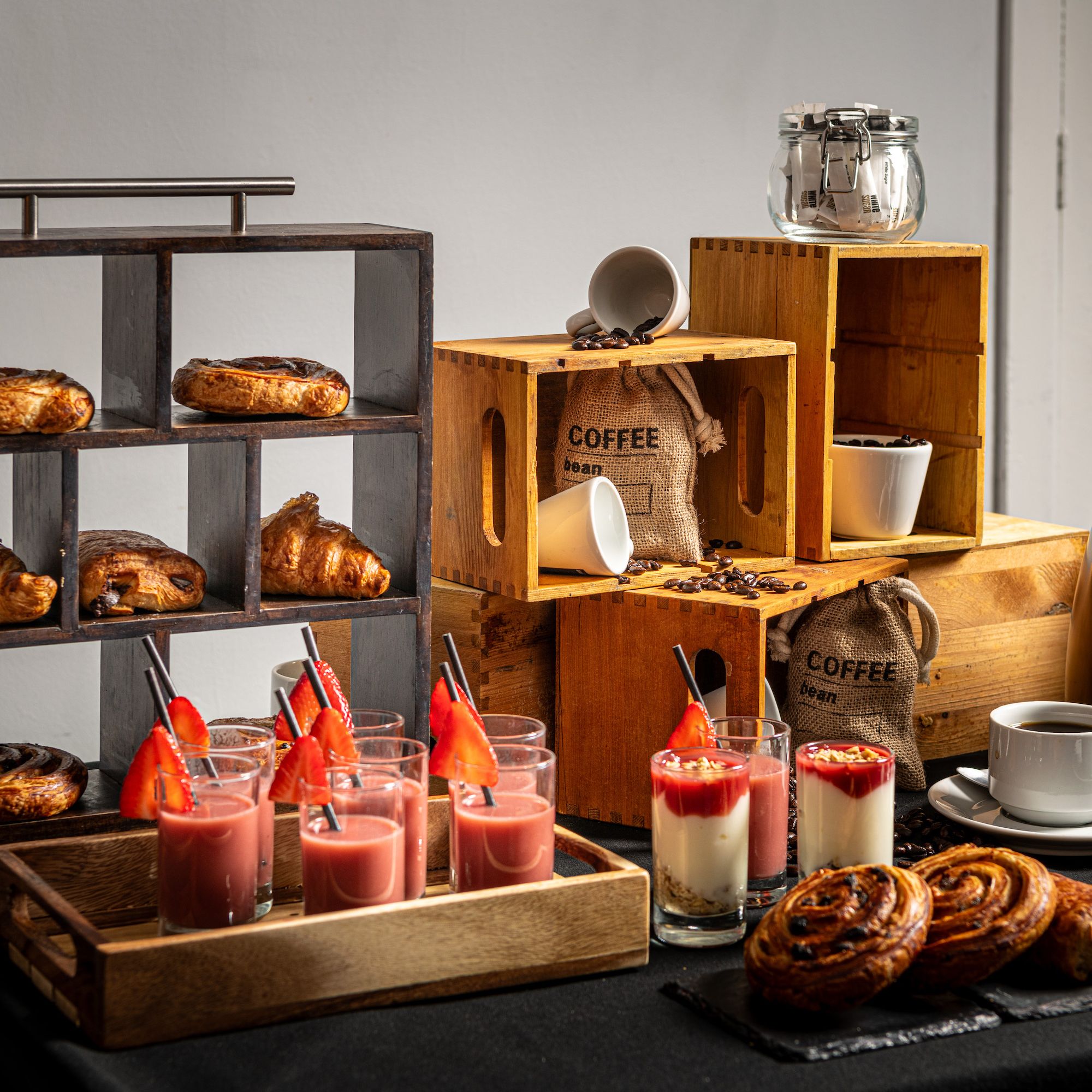 Breakfast buffet table with pastries, yogurt parfaits, coffee beans, cups, and juice glasses, arranged stylishly.