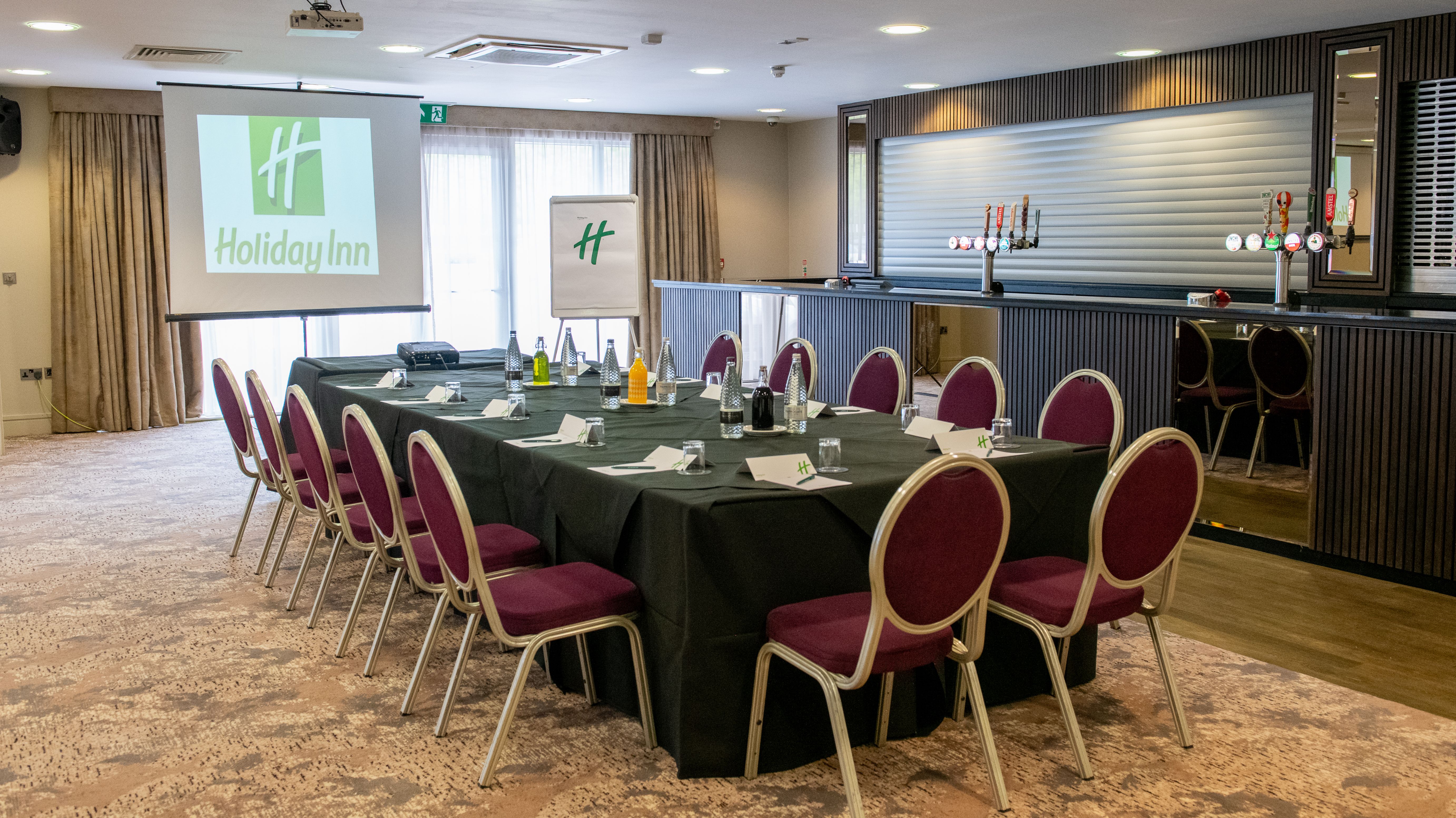 Conference room set up for a meeting at Holiday Inn with a large table, chairs, bottled water, and presentation screen.