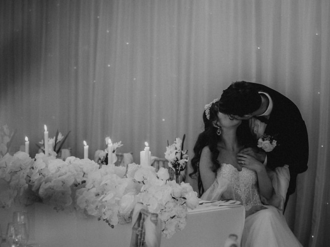 Bride and groom sharing a kiss at their wedding reception, surrounded by flowers and candles.