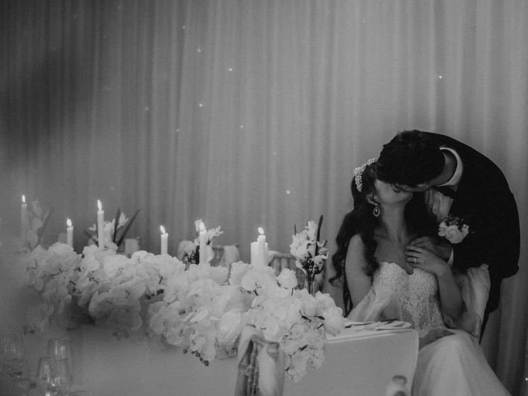 Bride and groom sharing a kiss at their wedding reception, surrounded by flowers and candles.