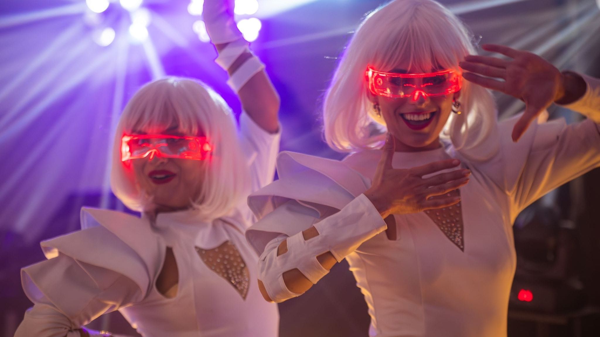 Two people in futuristic white costumes and wigs, wearing glowing red visor glasses, dancing under colorful stage lights.