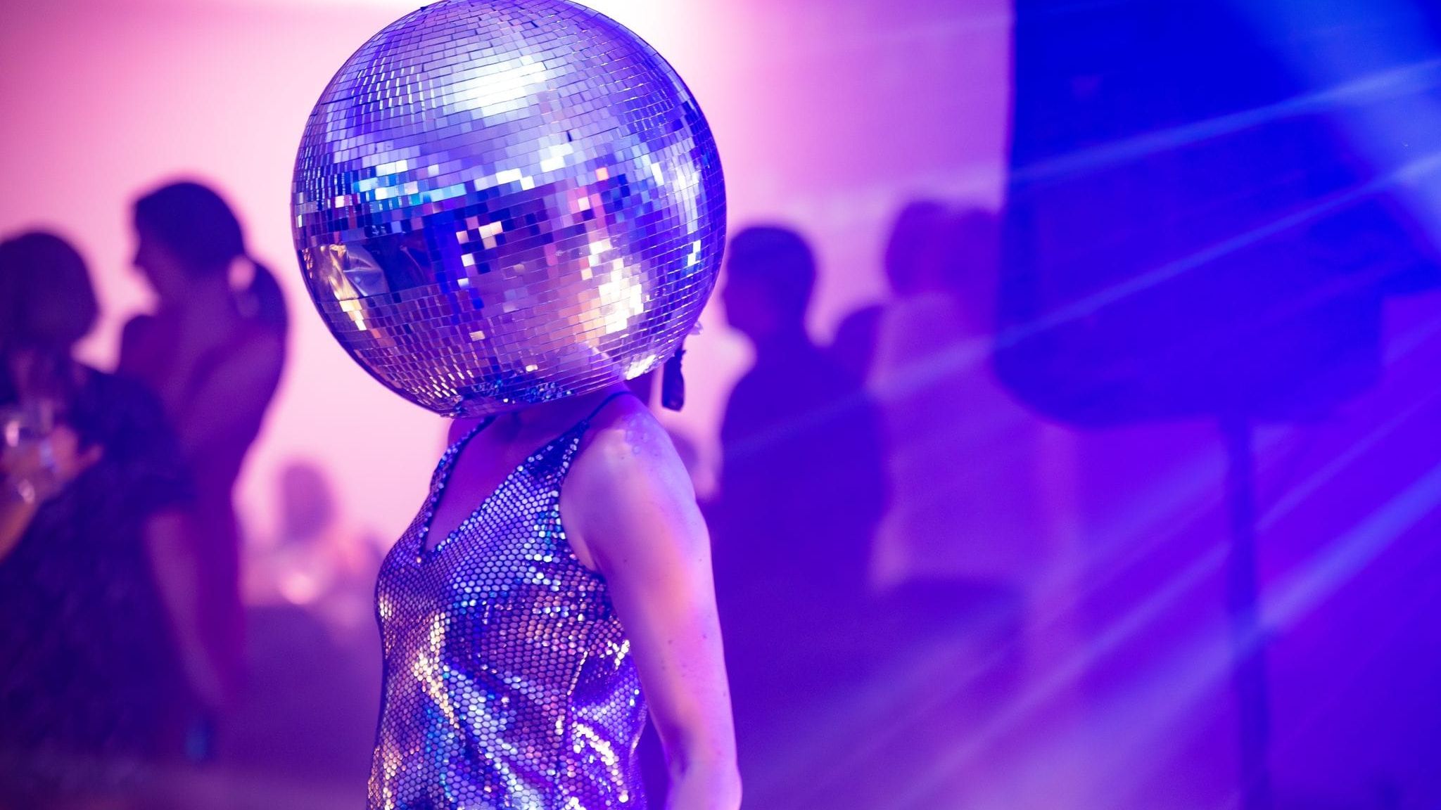 Person wearing a disco ball helmet and shiny outfit at a party with purple lighting