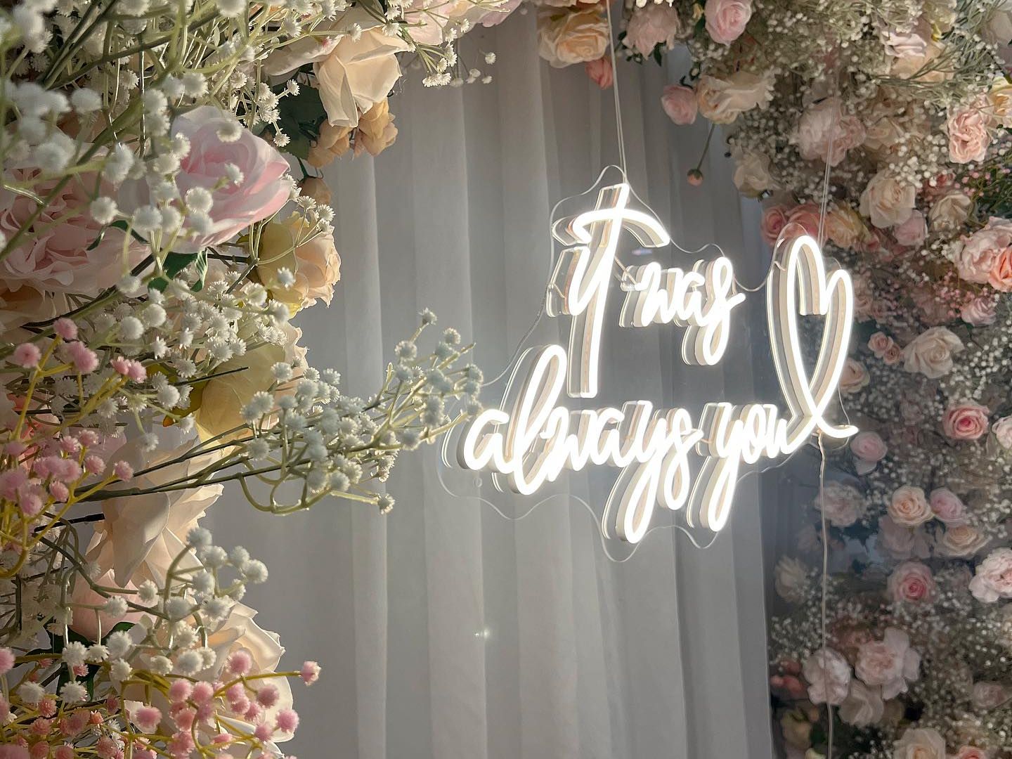 Neon sign reading 'It was always you' surrounded by pink and white flowers.