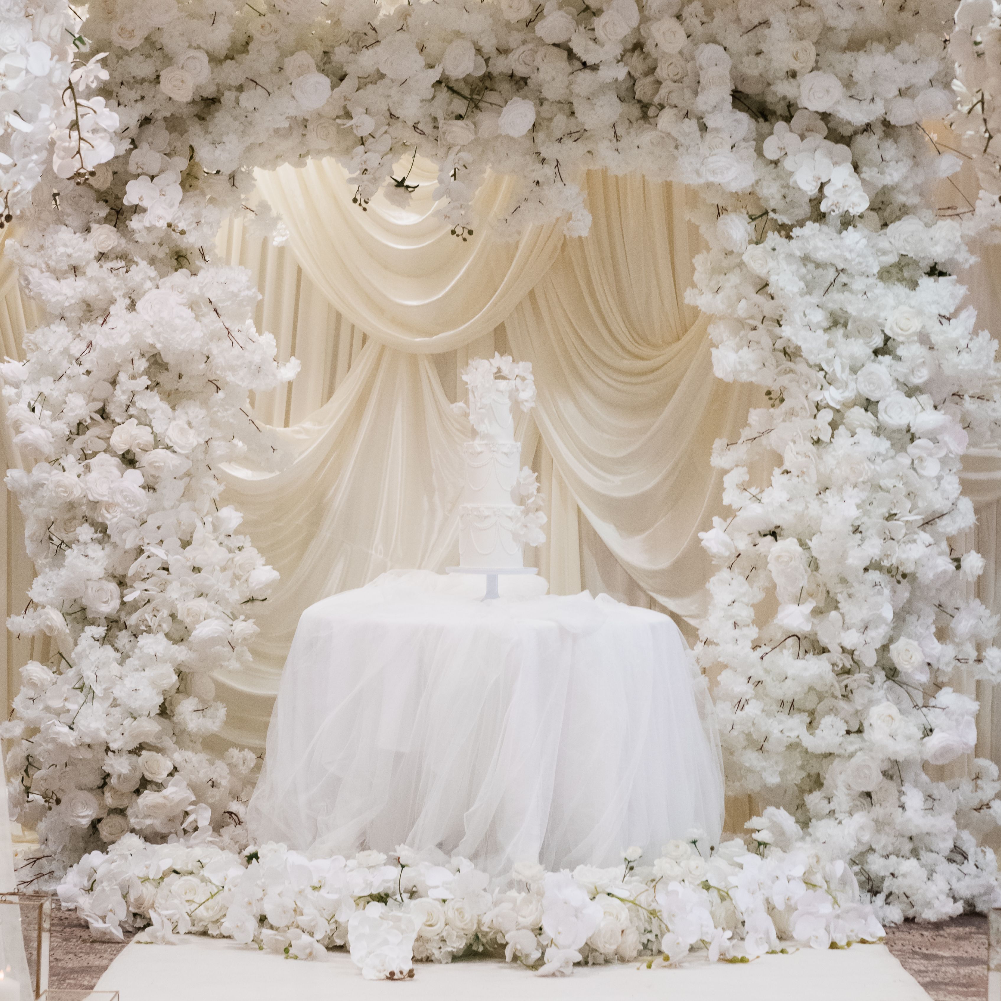 Elegant wedding cake display surrounded by white flowers and draped fabric backdrop