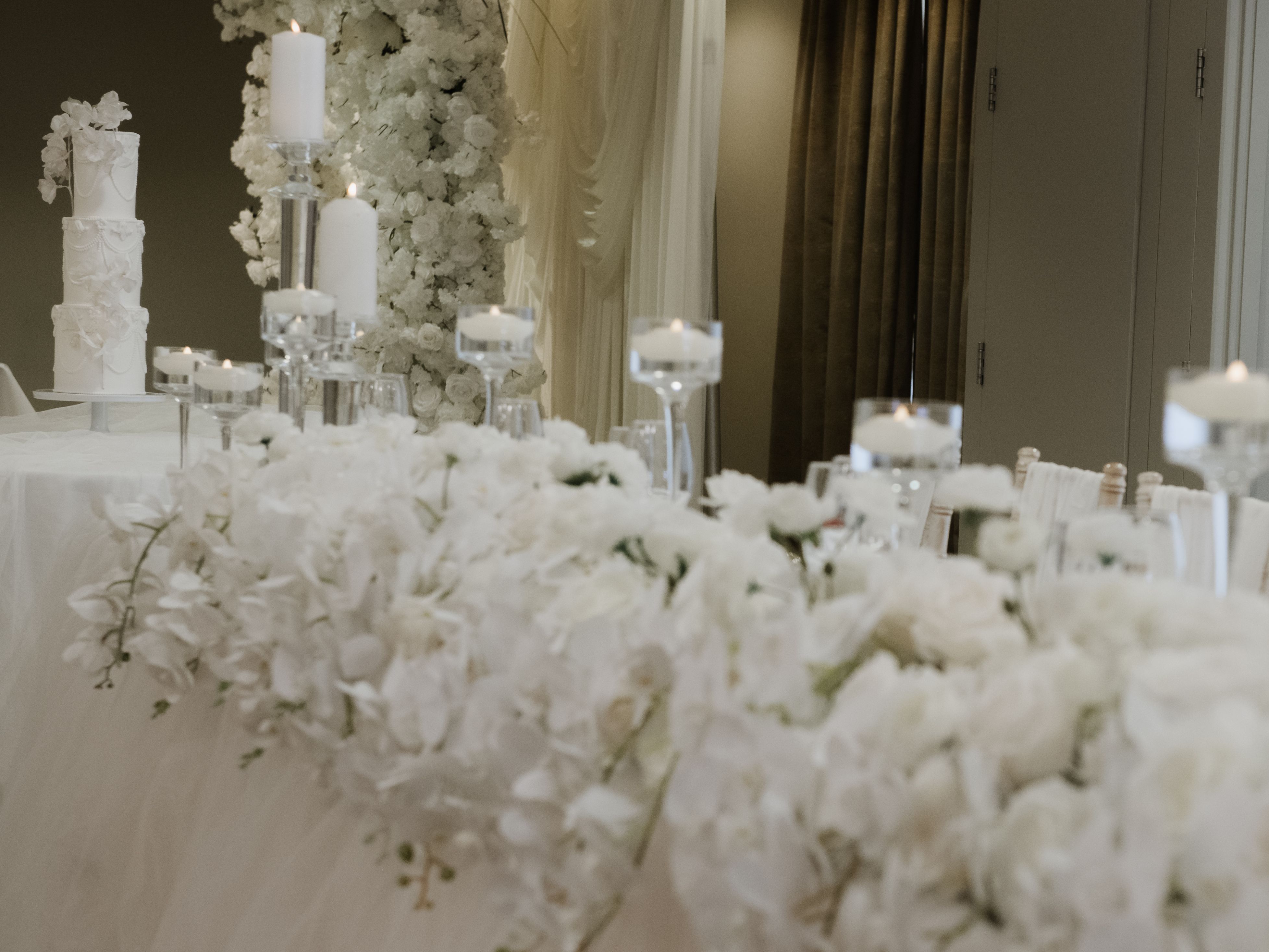 Elegant wedding reception table with white floral arrangements, candles, and a tiered white wedding cake in the background.