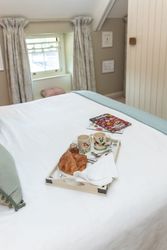 Breakfast tray with croissants, mugs, and jam on a bed in a cozy bedroom with a window and magazines.