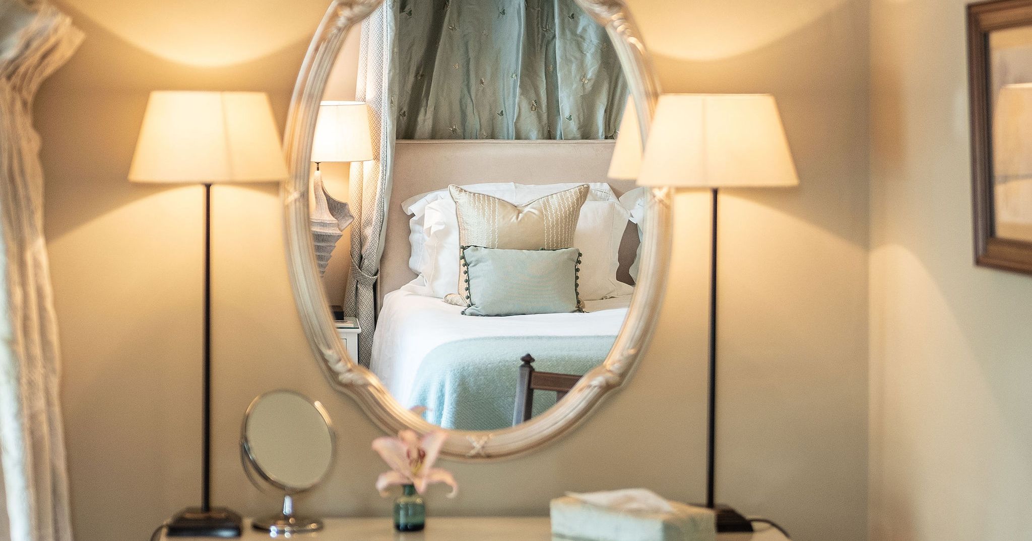 Elegant vanity table with lamps and chair, with a bed reflected in the oval mirror.