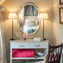 Elegant vanity table with lamps and chair, with a bed reflected in the oval mirror.