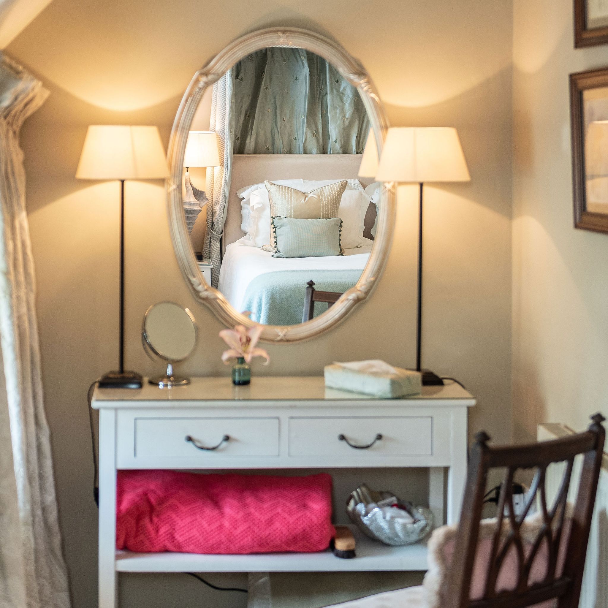 Elegant vanity table with lamps and chair, with a bed reflected in the oval mirror.