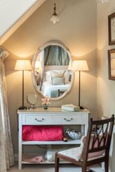 Elegant vanity table with lamps and chair, with a bed reflected in the oval mirror.