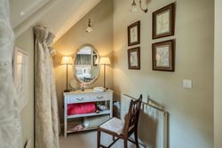 Cozy corner dressing table with an oval mirror, lamps, cushioned chair, and framed botanical art on pastel walls.