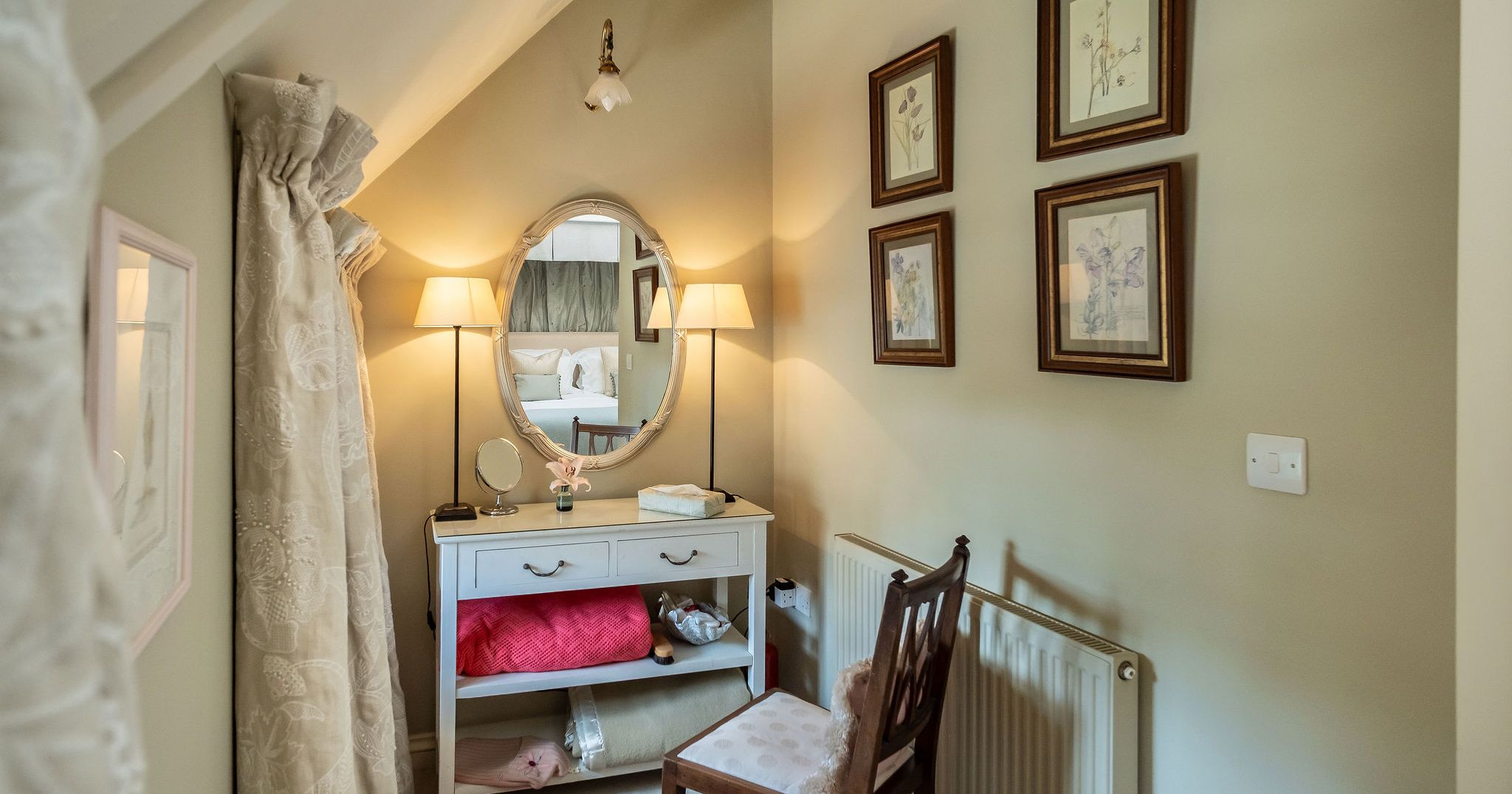 Cozy corner dressing table with an oval mirror, lamps, cushioned chair, and framed botanical art on pastel walls.