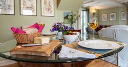 A cozy dining setup with bread, cheese, a bowl of salad, two glasses of white wine, and a small flower arrangement on a glass table in a warmly decorated living room.