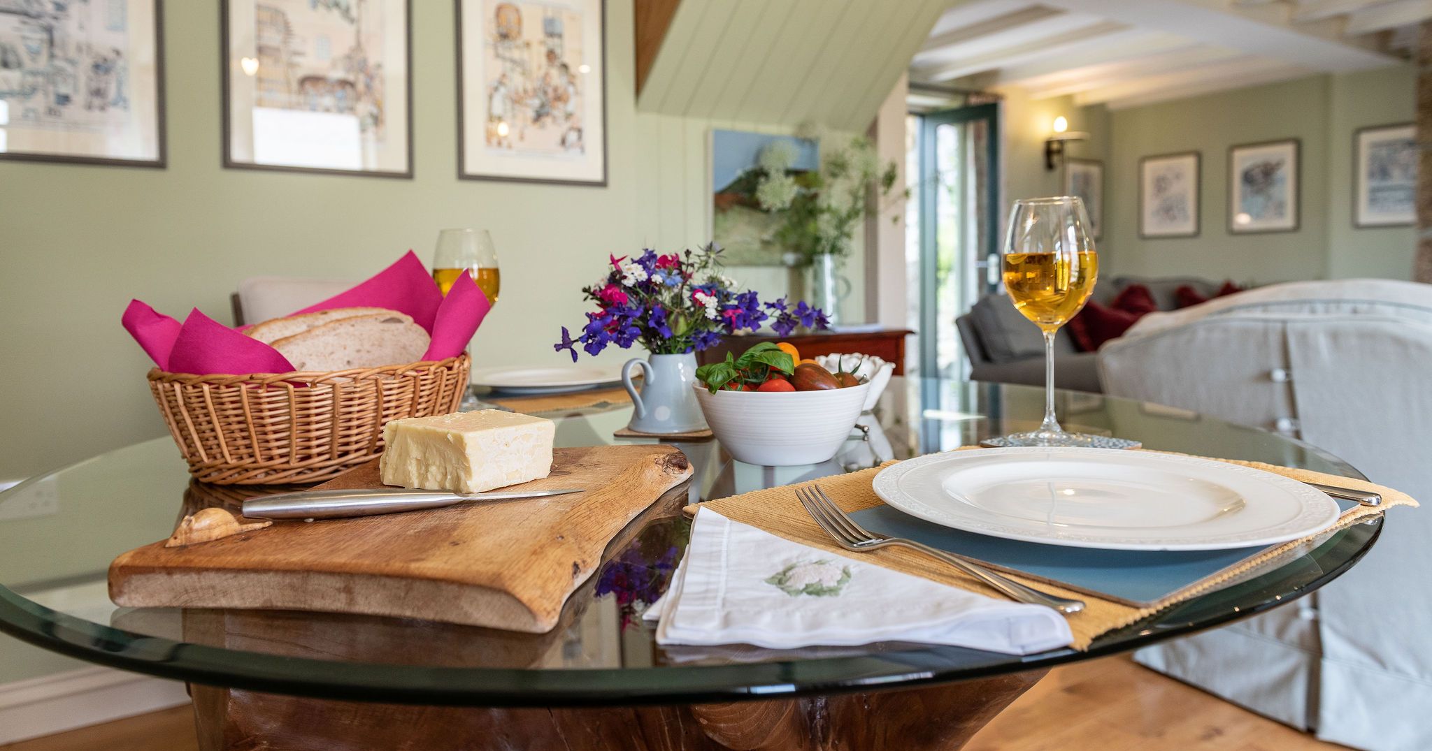 A cozy dining setup with bread, cheese, a bowl of salad, two glasses of white wine, and a small flower arrangement on a glass table in a warmly decorated living room.