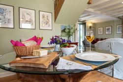 A cozy dining setup with bread, cheese, a bowl of salad, two glasses of white wine, and a small flower arrangement on a glass table in a warmly decorated living room.
