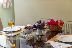 Glass dining table set for two with wine glasses, plates, a bowl of cherry tomatoes, a vase of fresh flowers, and a basket with pink napkins.