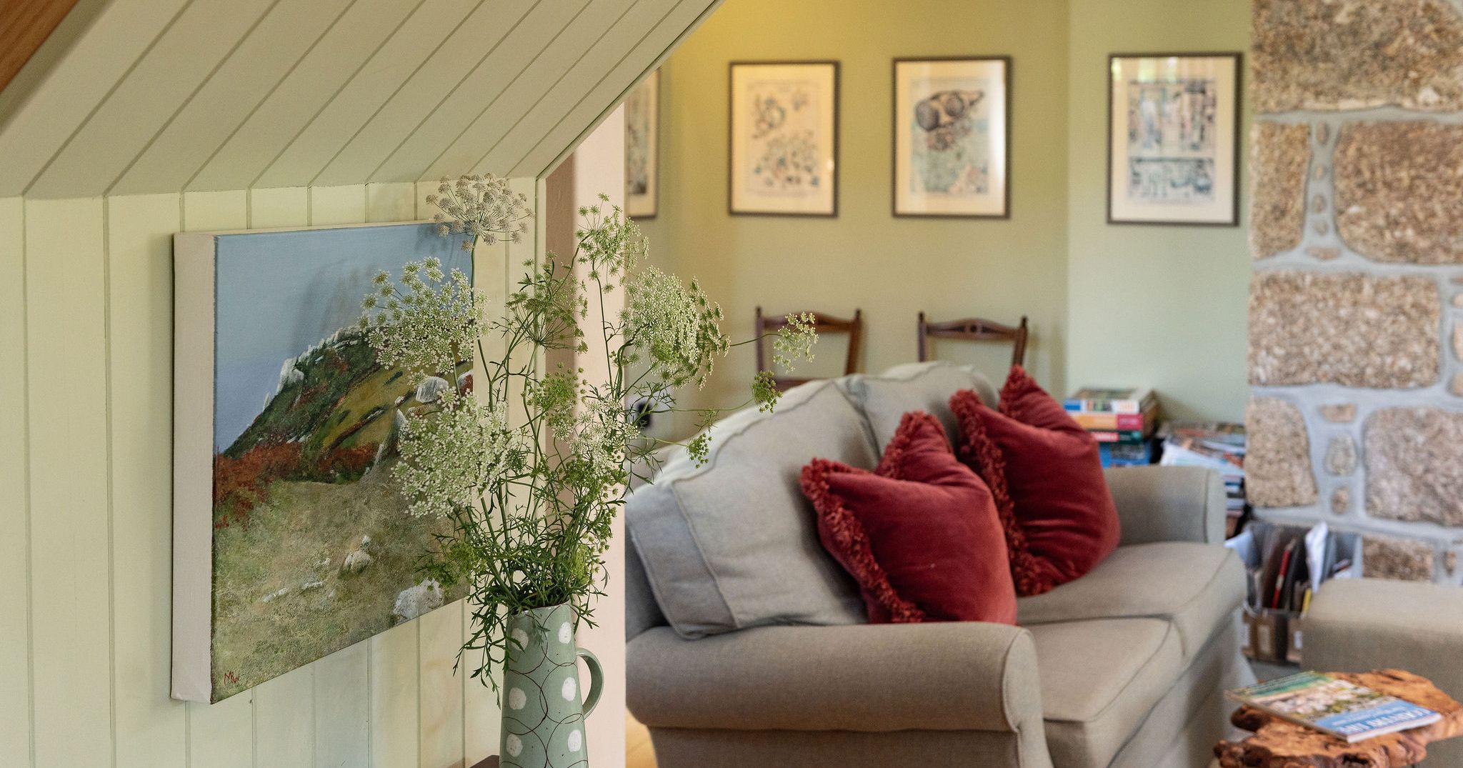 Cozy living room with a comfortable sofa, red cushions, framed artwork on the walls, and a vase of wildflowers on a wooden table.