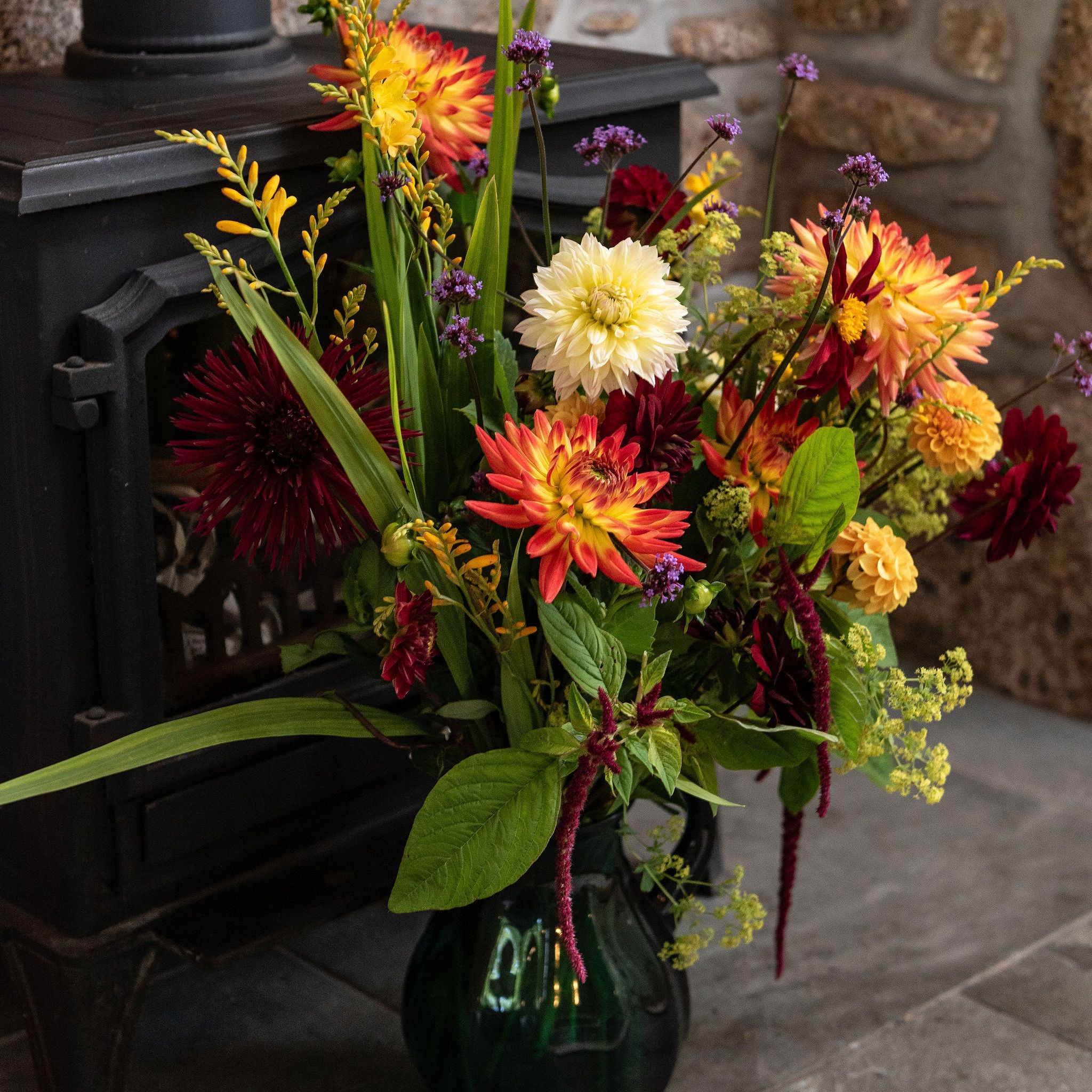 Colorful flower arrangement in a green vase placed next to a black wood-burning stove on a stone floor.