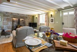 Cozy cottage living room with stone fireplace, soft armchairs, and a dining table set with wine, cheese, and flowers.