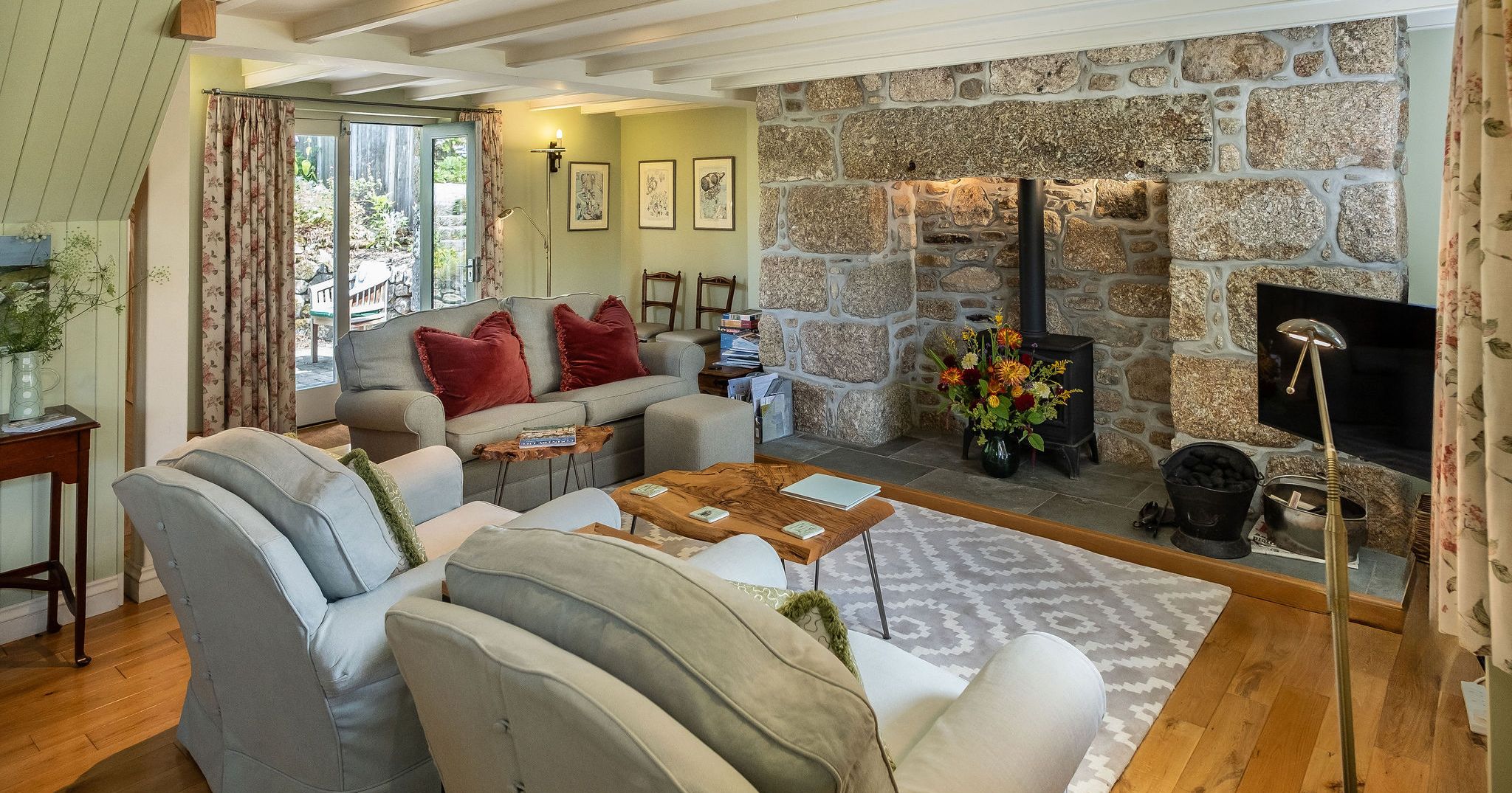 Cozy cottage living room with stone fireplace, wood-burning stove, comfortable armchairs, and a sofa with red pillows.