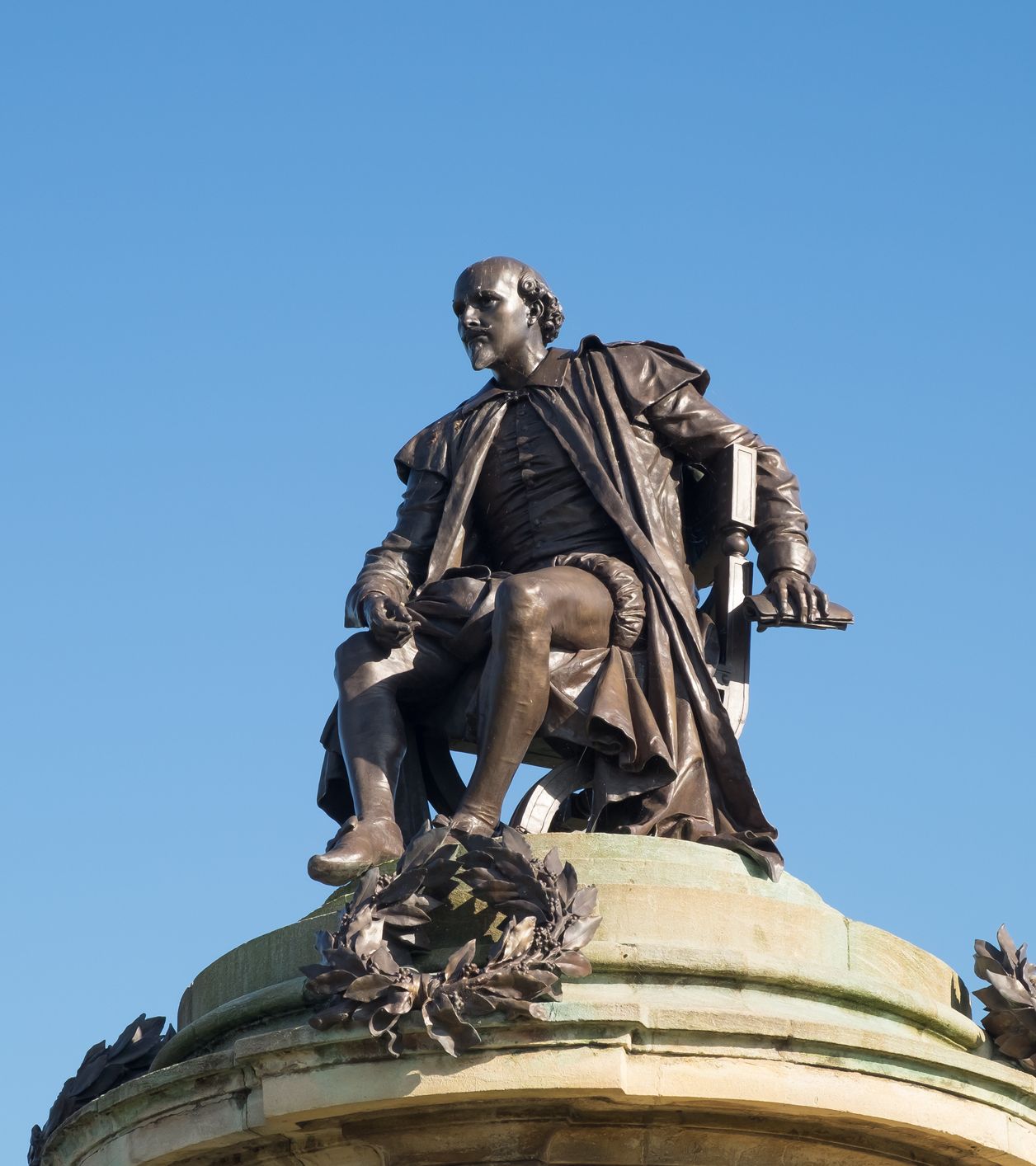Statue of William Shakespeare seated atop a monument, gazing thoughtfully into the distance, with a laurel wreath below, set against a clear blue sky.
