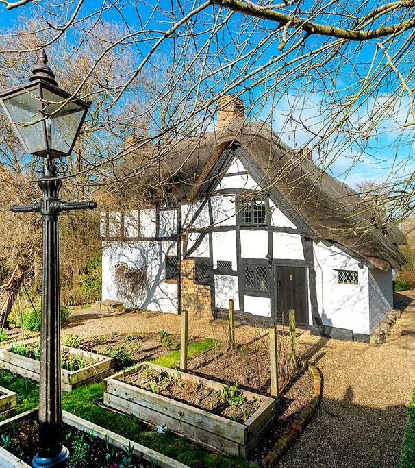 Charming thatched-roof cottage with black timber framing, surrounded by garden beds, a stone pathway, and an old-fashioned street lamp under clear blue skies.