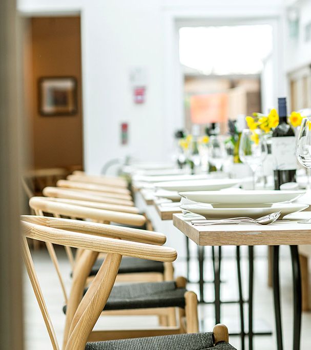 Bright, modern restaurant with a row of neatly set tables featuring wine bottles, glasses, and fresh flowers. Light wood chairs add a natural, elegant touch.