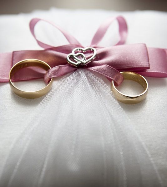 Close-up of two gold wedding rings on a white satin pillow, adorned with a pink ribbon and silver heart decoration, symbolizing love and commitment.