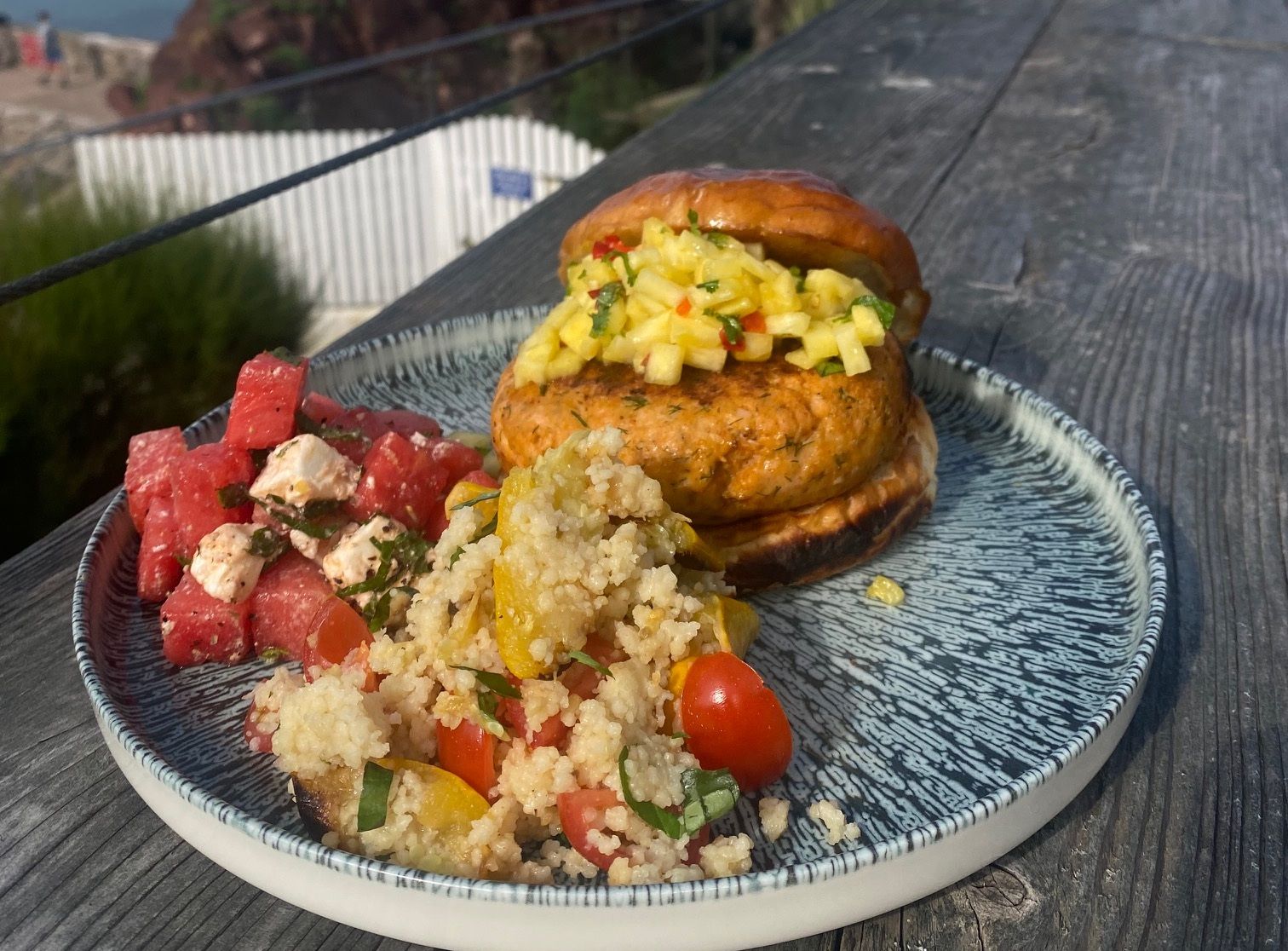 A plate with a salmon burger topped with pineapple salsa, a watermelon feta salad, and a couscous salad, set on a wooden table overlooking the ocean and a rocky outcrop.
