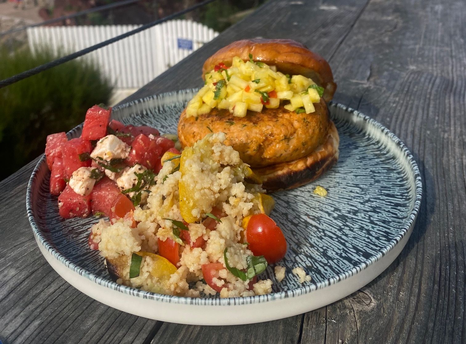 A plate with a salmon burger topped with pineapple salsa, a watermelon feta salad, and a couscous salad, set on a wooden table overlooking the ocean and a rocky outcrop.