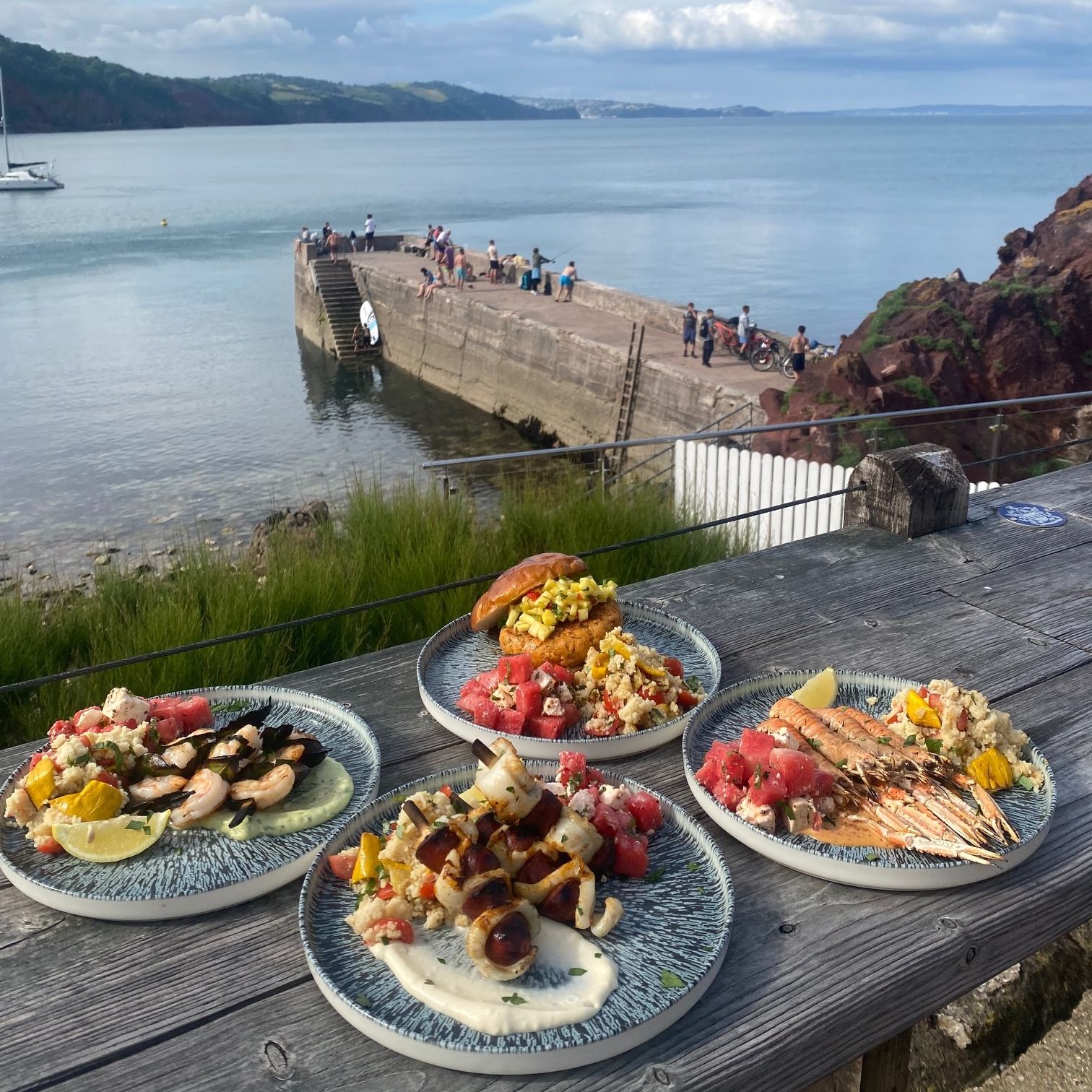 Plates of seafood and salad on a wooden table overlooking a harbor with people standing on a pier and a sailboat on the water.