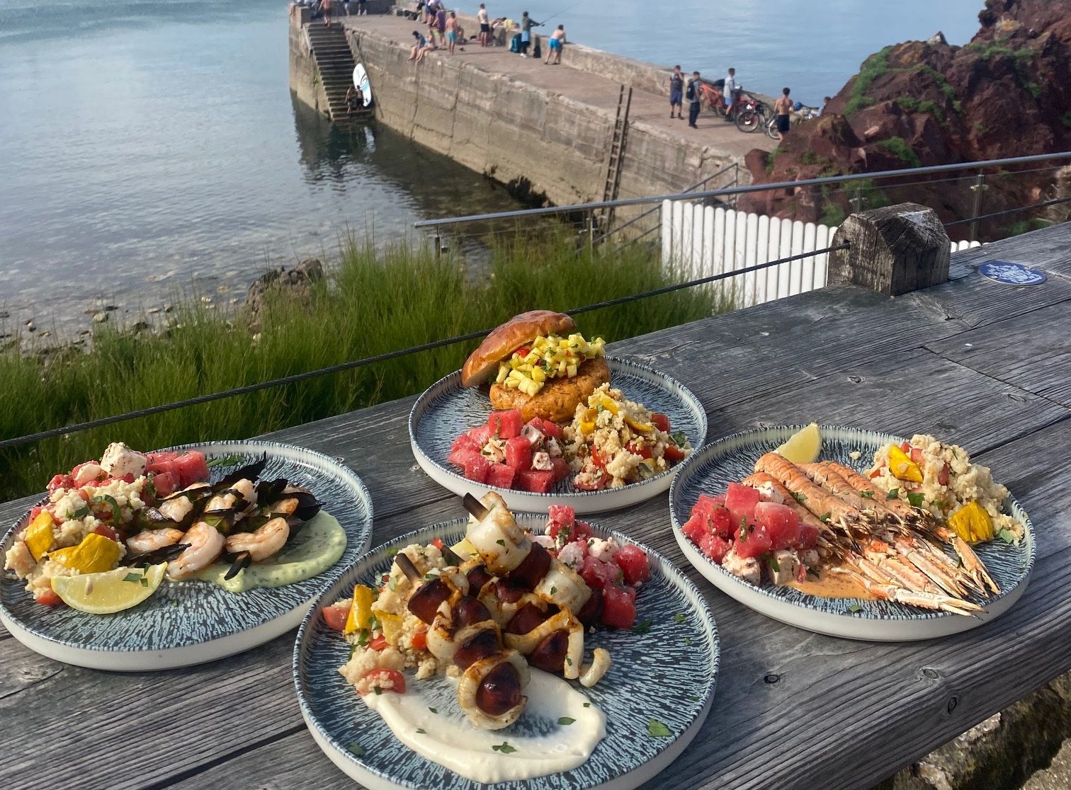 Plates of seafood and salad on a wooden table overlooking a harbor with people standing on a pier and a sailboat on the water.