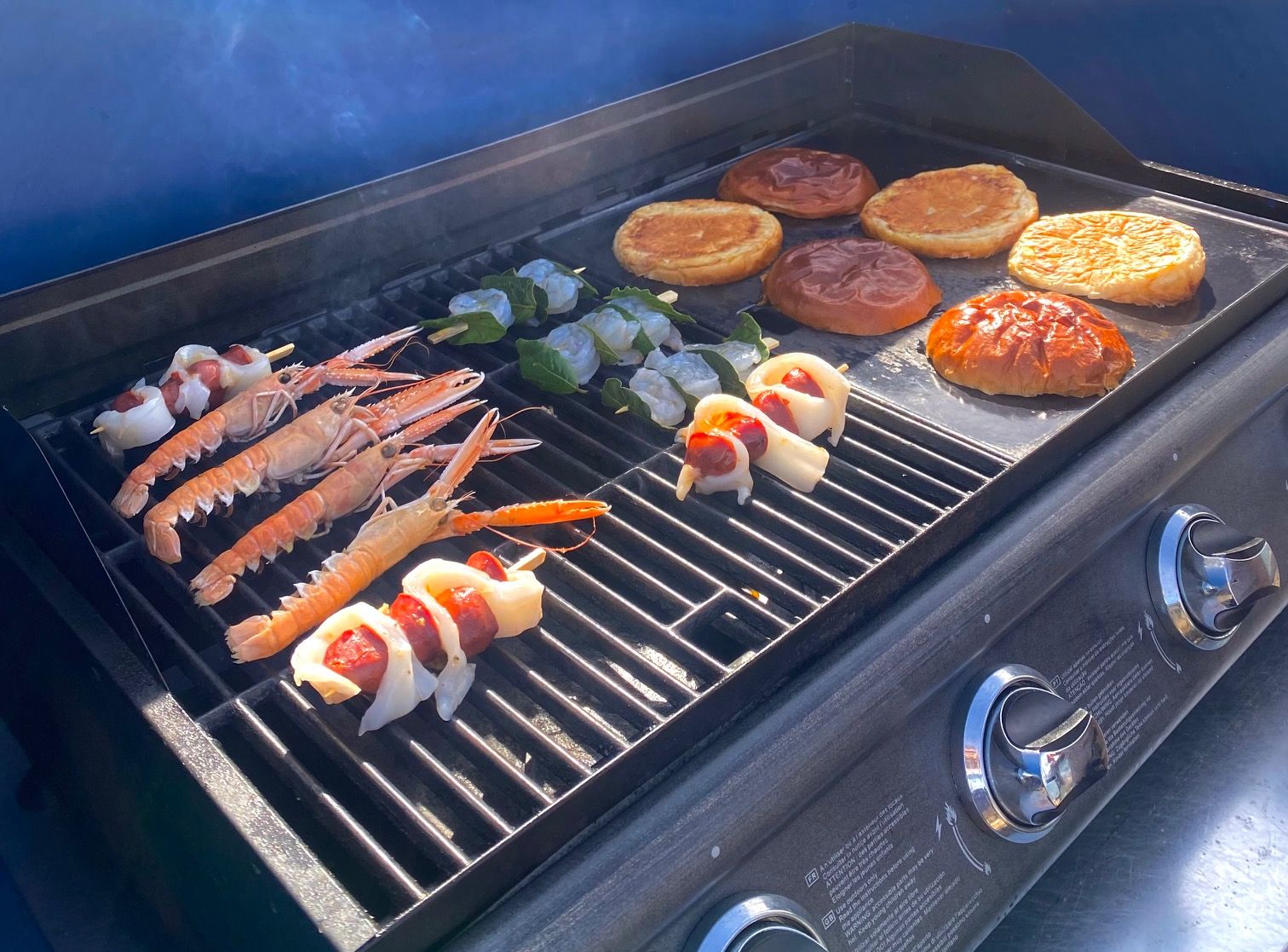 Seafood, bread, and vegetable skewers being grilled on an outdoor barbecue under sunlight.