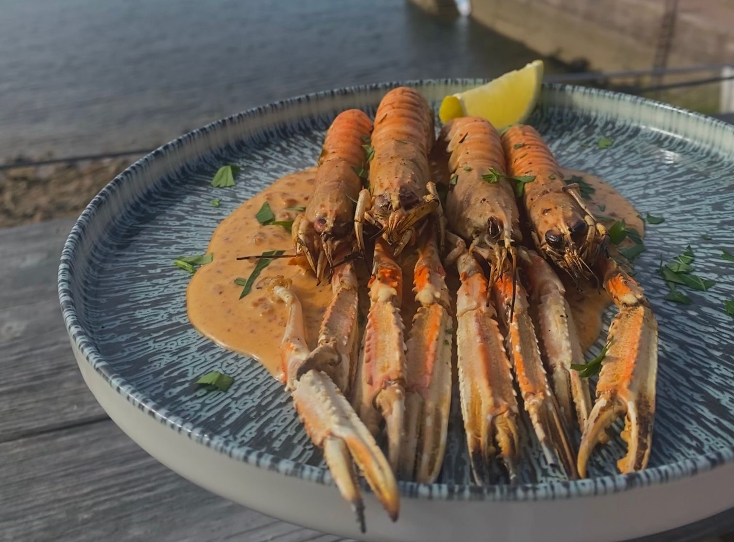 Plate of langoustines with sauce, served outdoors by the sea with a view of a harbor and sailboat.