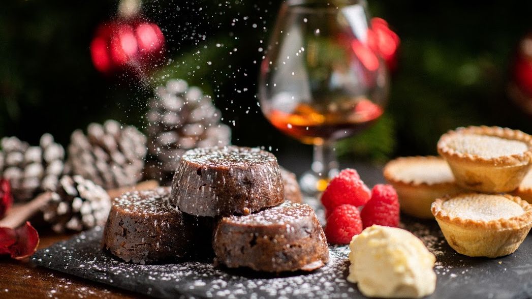 Christmas desserts with chocolate lava cakes, mince pies, raspberries, cream, and a glass of brandy with festive decorations in the background.
