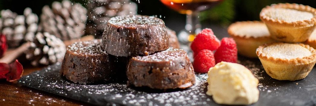 Christmas desserts with chocolate lava cakes, mince pies, raspberries, cream, and a glass of brandy with festive decorations in the background.