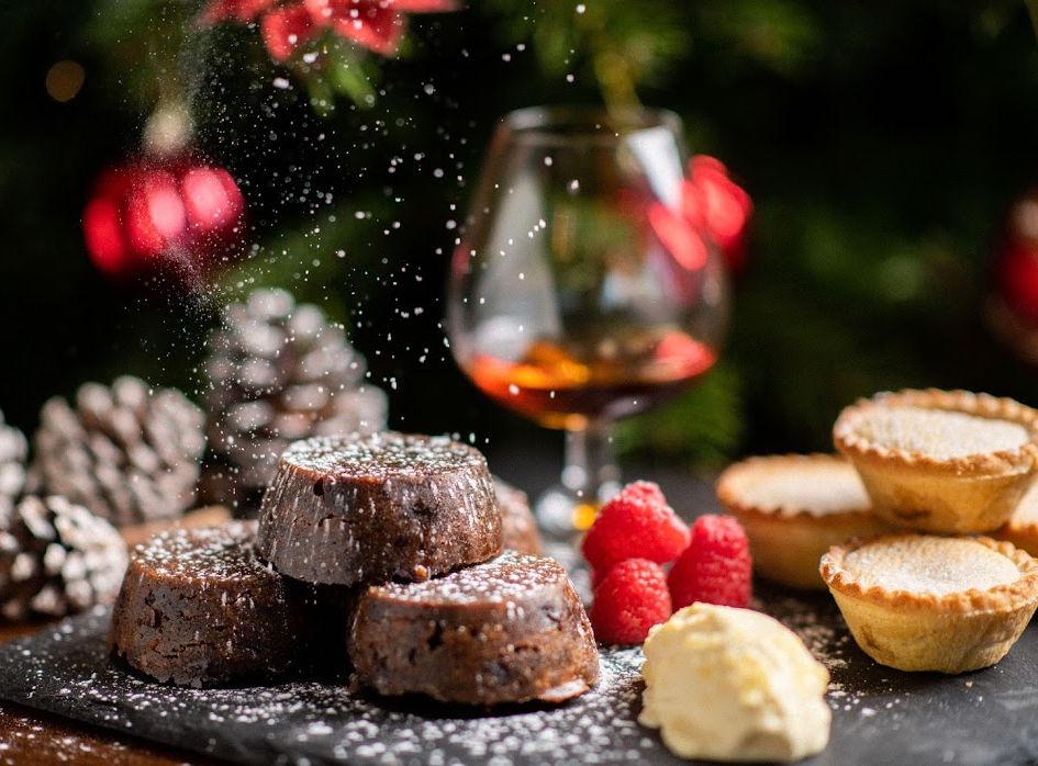 Christmas desserts with chocolate lava cakes, mince pies, raspberries, cream, and a glass of brandy with festive decorations in the background.