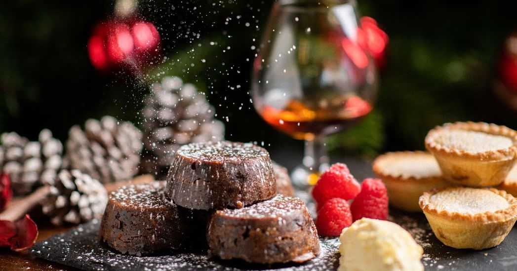 Christmas desserts with chocolate lava cakes, mince pies, raspberries, cream, and a glass of brandy with festive decorations in the background.