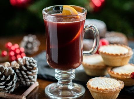 Glass of mulled wine with a lemon slice, mince pies, pine cones, and Christmas tree in the background