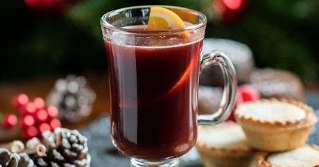 Glass of mulled wine with a lemon slice, mince pies, pine cones, and Christmas tree in the background