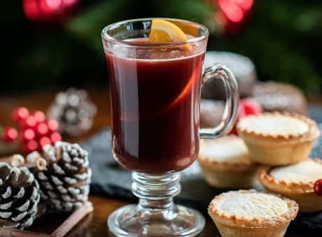 Glass of mulled wine with a lemon slice, mince pies, pine cones, and Christmas tree in the background