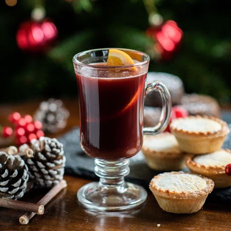 Glass of mulled wine with a lemon slice, mince pies, pine cones, and Christmas tree in the background
