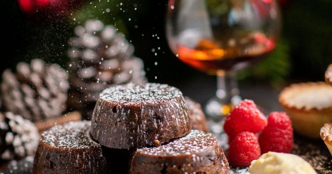 Festive chocolate desserts with powdered sugar, raspberries, cream, a glass of brandy, pine cones, and Christmas decorations in the background.