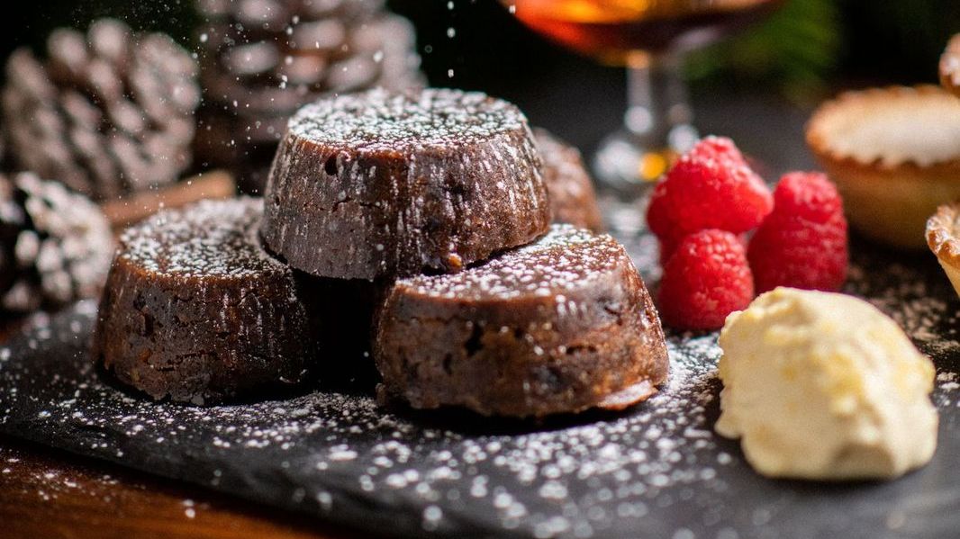 Festive chocolate desserts with powdered sugar, raspberries, cream, a glass of brandy, pine cones, and Christmas decorations in the background.