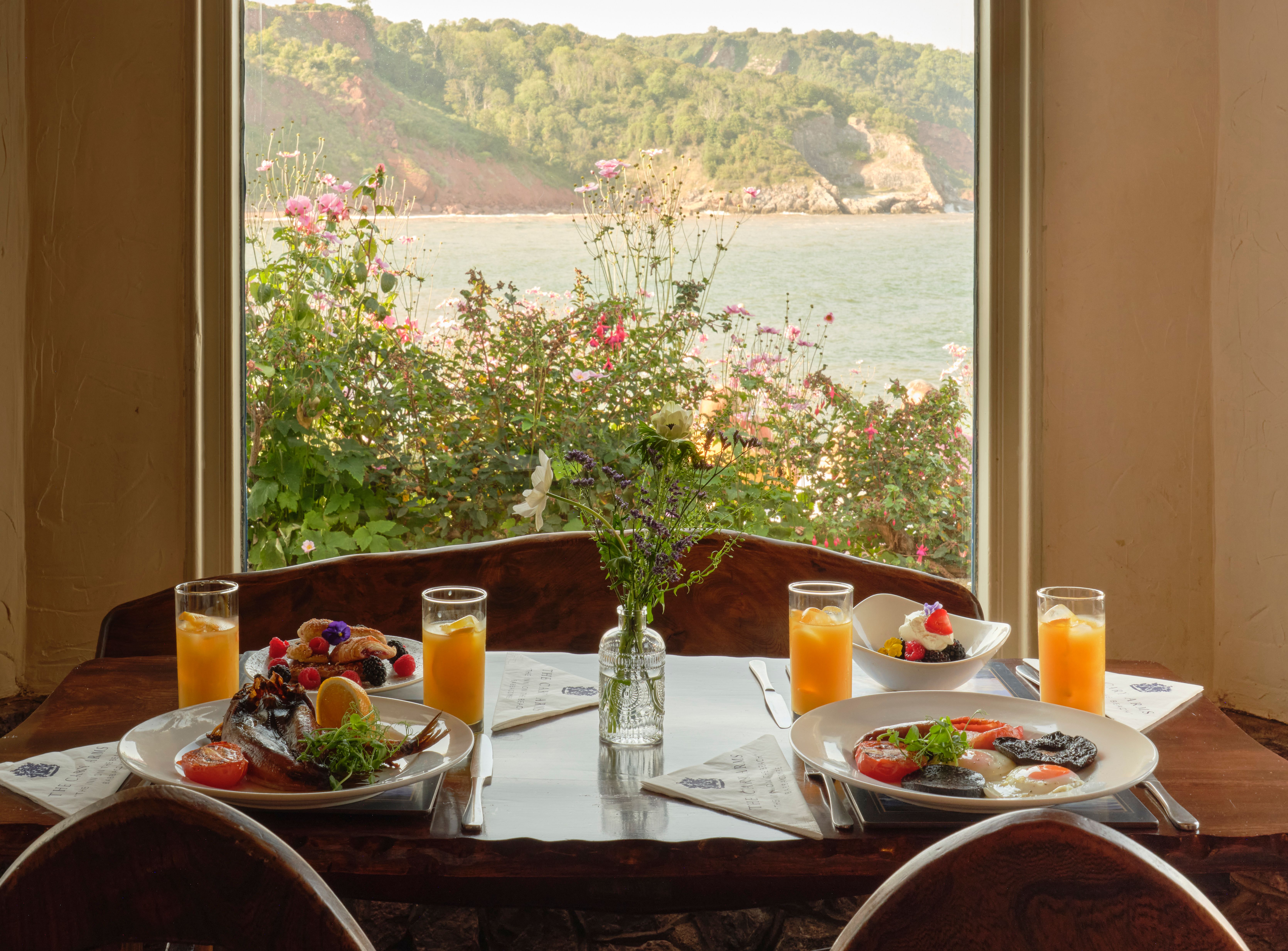 Breakfast table with plates of food and orange juice by a window overlooking the sea and cliffs