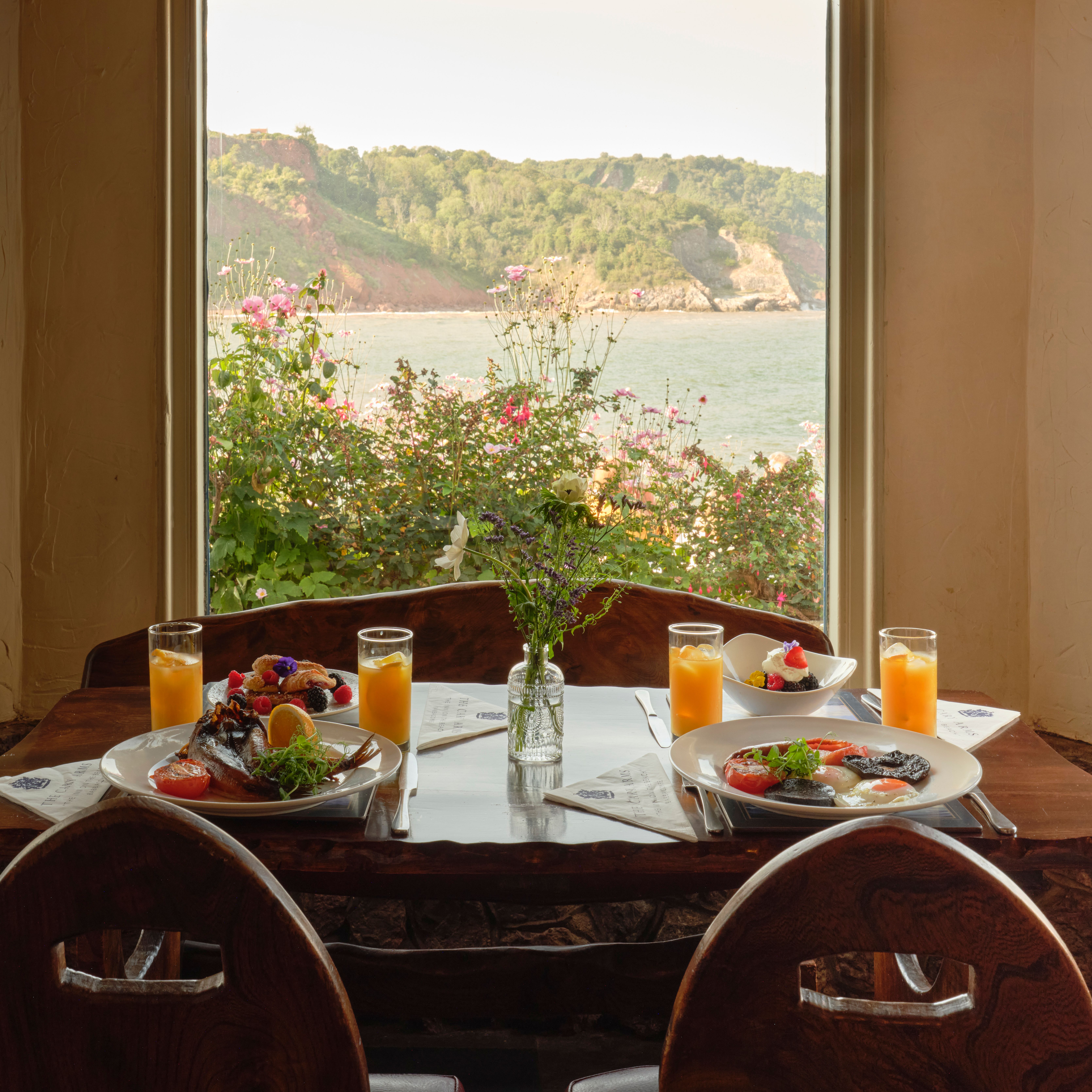 Breakfast table with plates of food and orange juice by a window overlooking the sea and cliffs