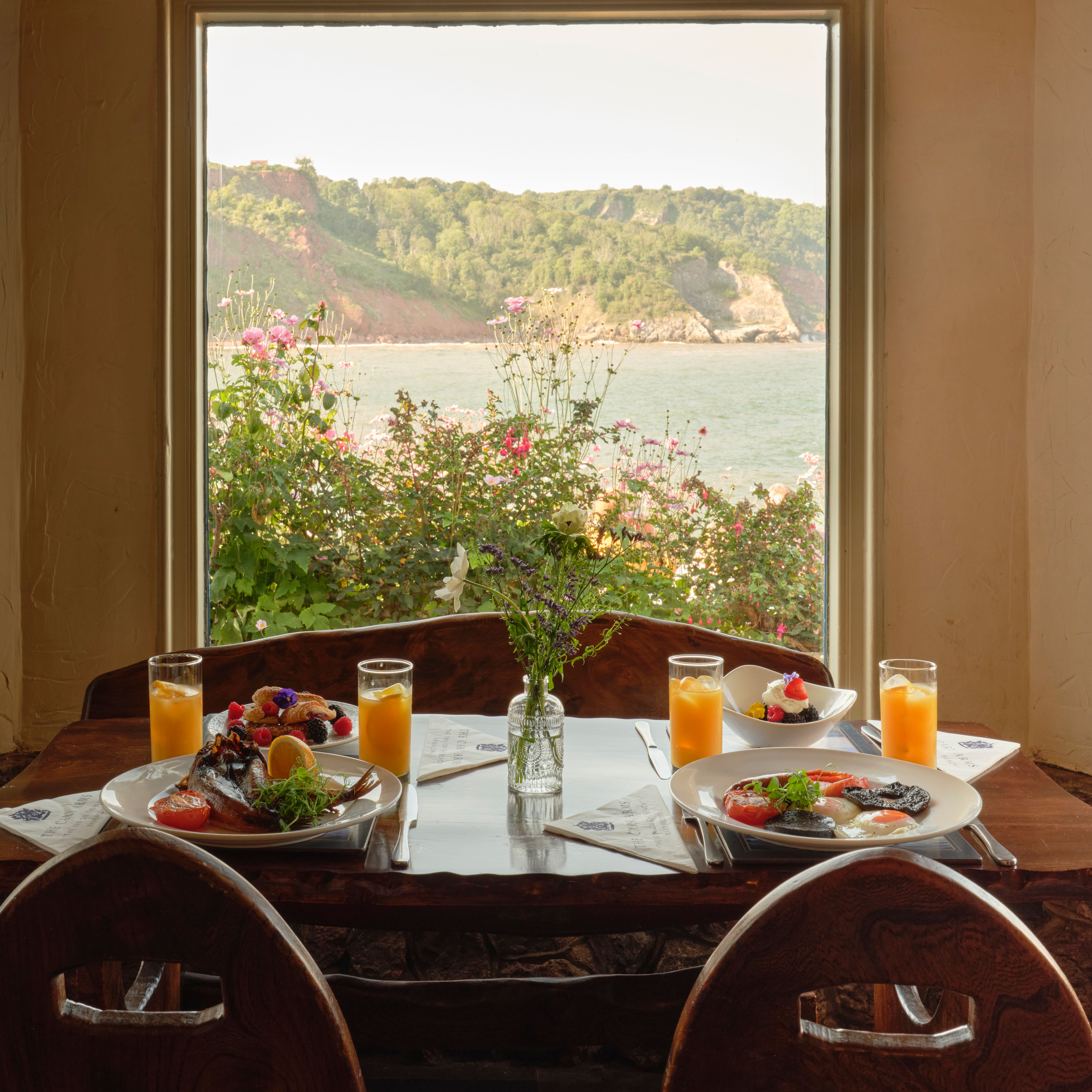 Breakfast table with plates of food and orange juice by a window overlooking the sea and cliffs