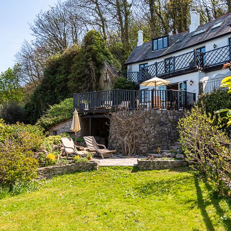 Charming countryside house with a stone terrace, patio chairs, and lush green garden surrounded by trees and bushes on a sunny day.
