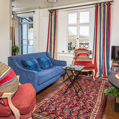 Cozy living room with a blue sofa, red armchair, patterned cushions, ornate rug, striped curtains, and small table, decorated with gold accents and houseplants.